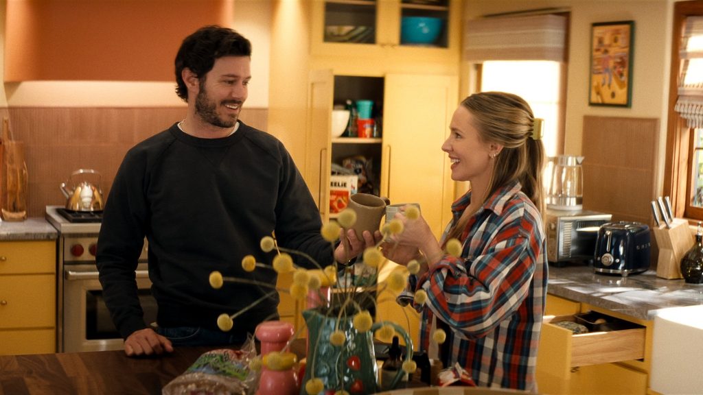 "A man and woman smile and toast with mugs in a cozy, sunlit kitchen, surrounded by yellow cabinets, kitchen appliances, and decorative items, creating a warm, homey atmosphere."