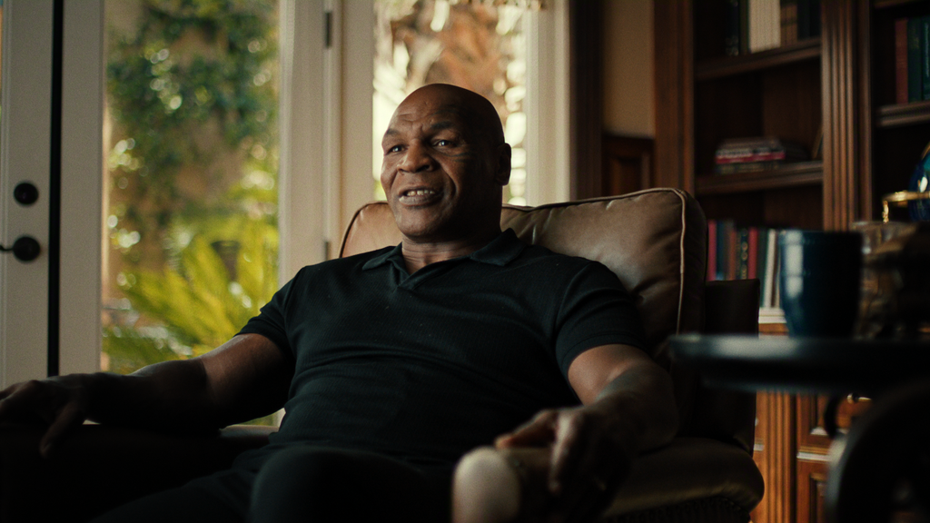 "A man sitting in a cozy armchair in a warmly lit home, surrounded by bookshelves and large windows with sunlight and plants visible outside, a coffee table with a mug in the foreground."