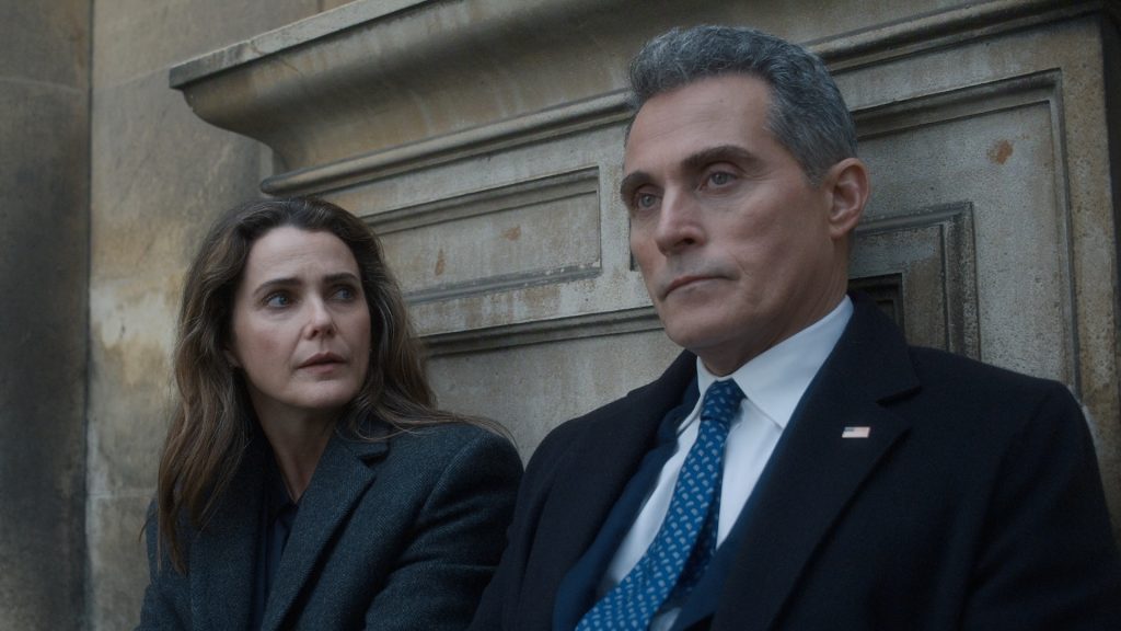 "Two people in formal coats sit on outdoor stone steps, appearing serious and deep in conversation, with an old stone wall in the background. The man wears a suit and tie with an American flag pin, suggesting a political or official context."